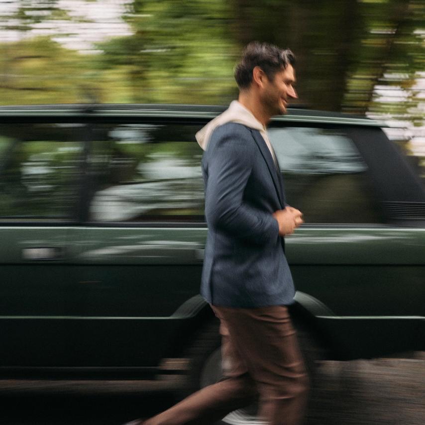 Man in a navy blazer layered over a cream hoodie with brown pants, striding past a green Range Rover