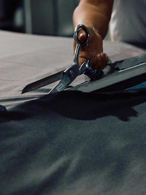 Close-up shot of a tailor's hands cutting grey fabric with large metal shears.