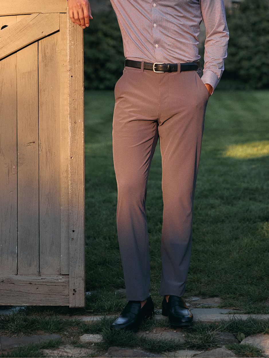 Lower-body shot of a man resting his hand against a wooden gate, wearing dark grey pants, a black belt, and black loafers.