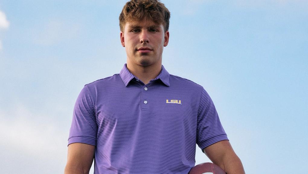 A man in a purple striped LSU polo shirt holding a football under his arm, standing outdoors against a mountain background.