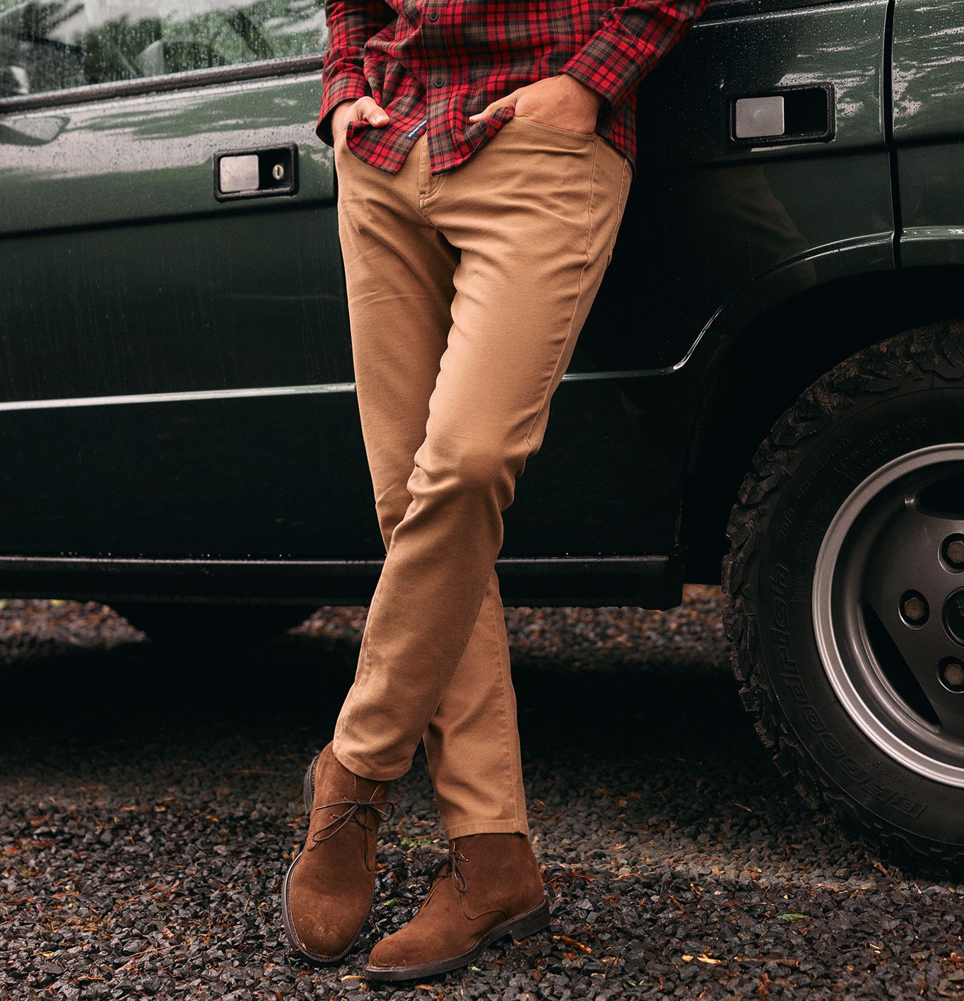 A close-up shot focusing on tan pants and brown suede boots worn by a man leaning against a green SUV.