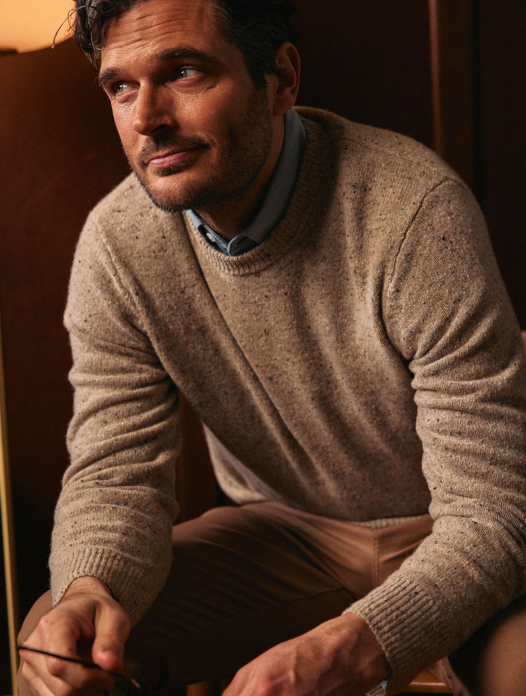 A man wearing a tan speckled crewneck sweater over a light blue collared shirt sitting indoors.