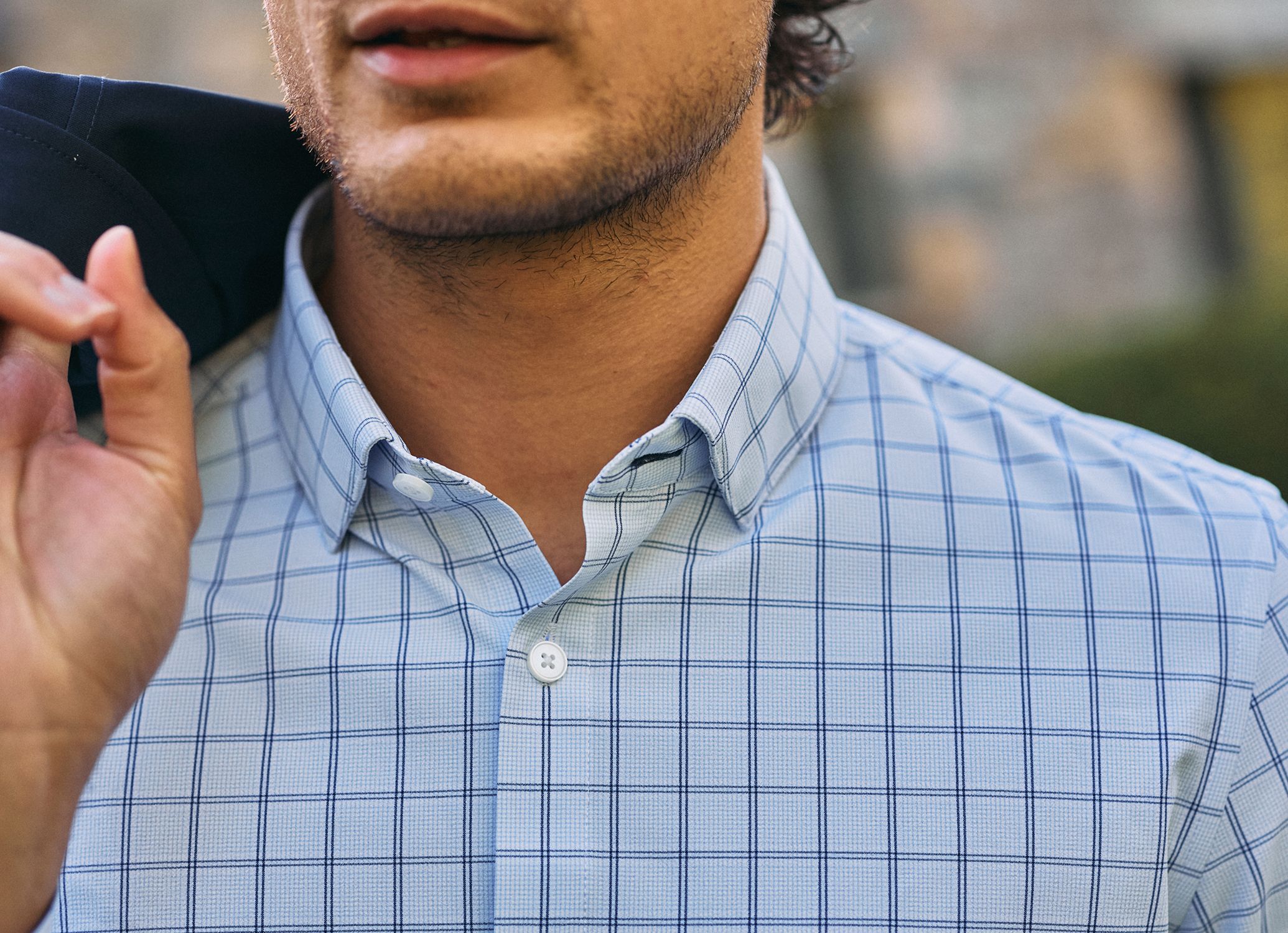 Close-up of a man in a light blue windowpane check dress shirt holding a navy jacket over his shoulder