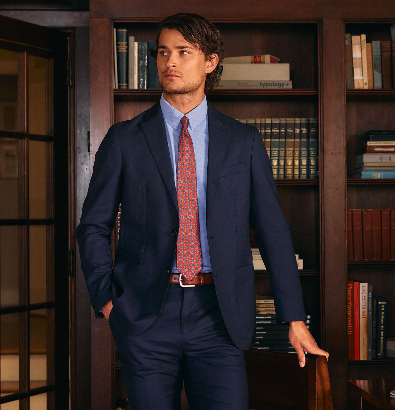 A man wearing a dark blue suit, light blue shirt, and red pattern tie standing in front of dark wooden bookshelves.