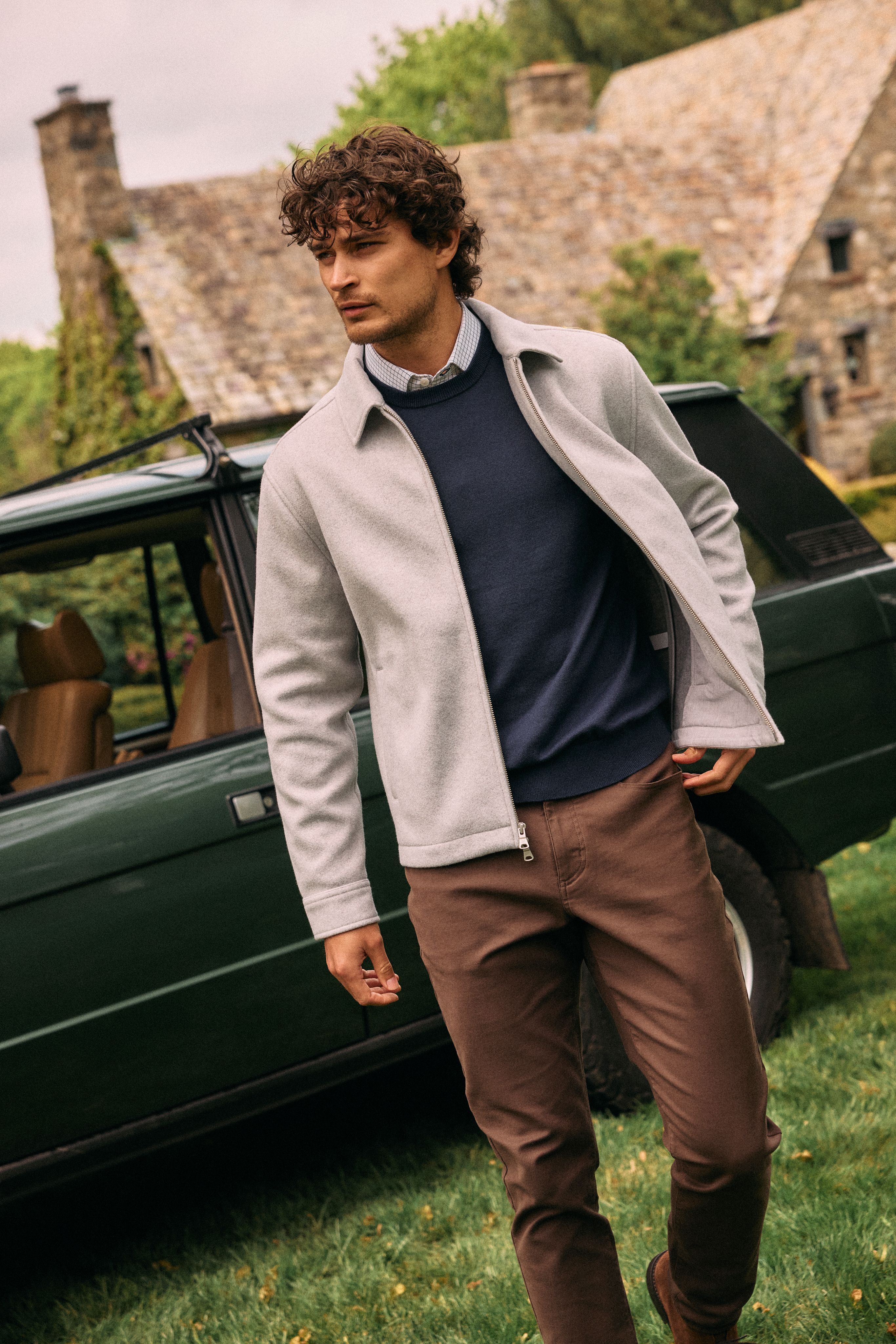 Man in a light gray jacket over a navy sweater and brown pants walking past a vintage green Range Rover