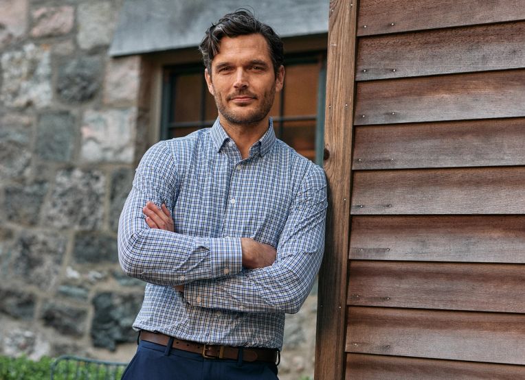 Man in a blue check dress shirt and navy pants leaning against a weathered wood wall with his arms crossed