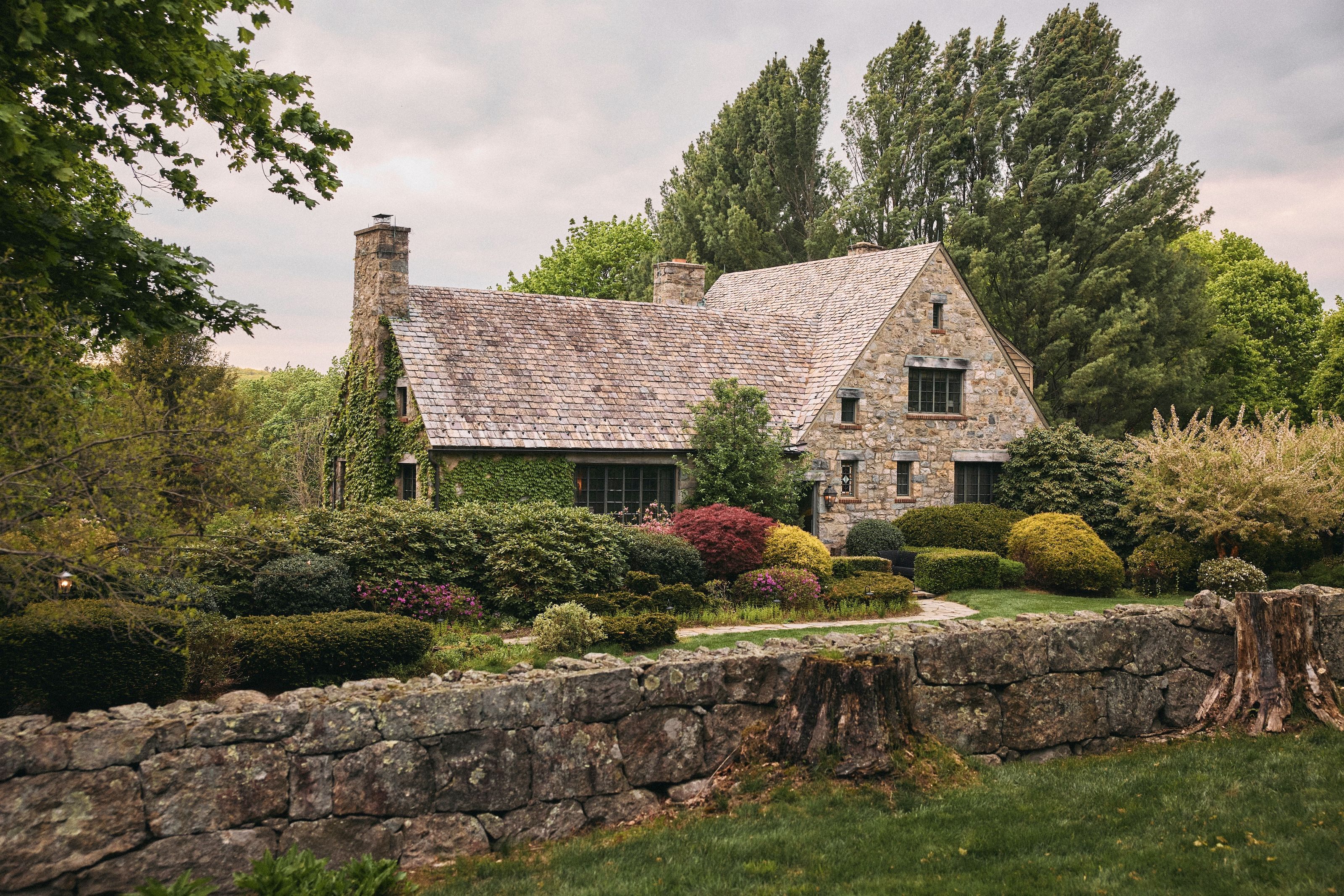 An ivy-covered stone country house in Connecticut behind a low stone garden wall, surrounded by lush gardens and overhanging branches.