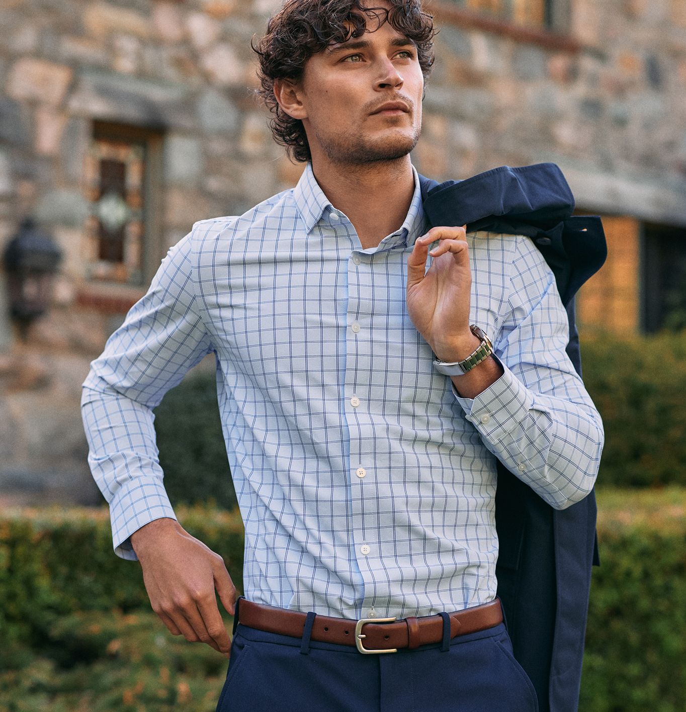 A man in a light blue grid-pattern button-down shirt holding a suit jacket over his shoulder while standing outdoors in front of a stone building.