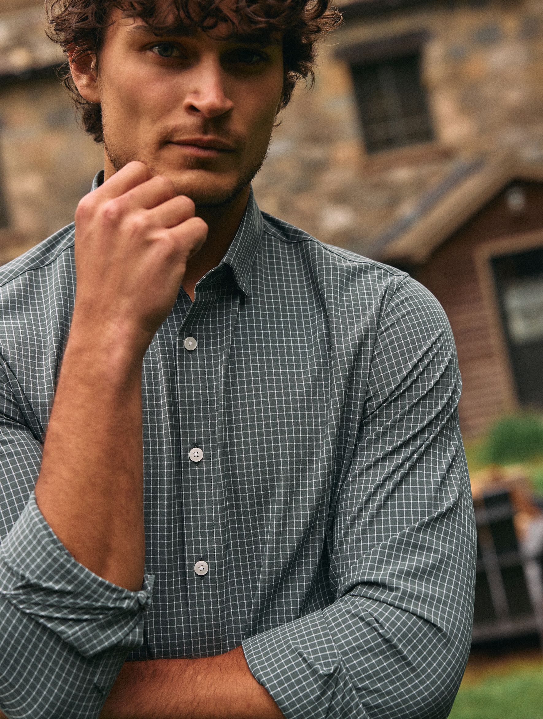 A close-up portrait of a man with his hand to his chin wearing a green and white grid-pattern button-down shirt outdoors.