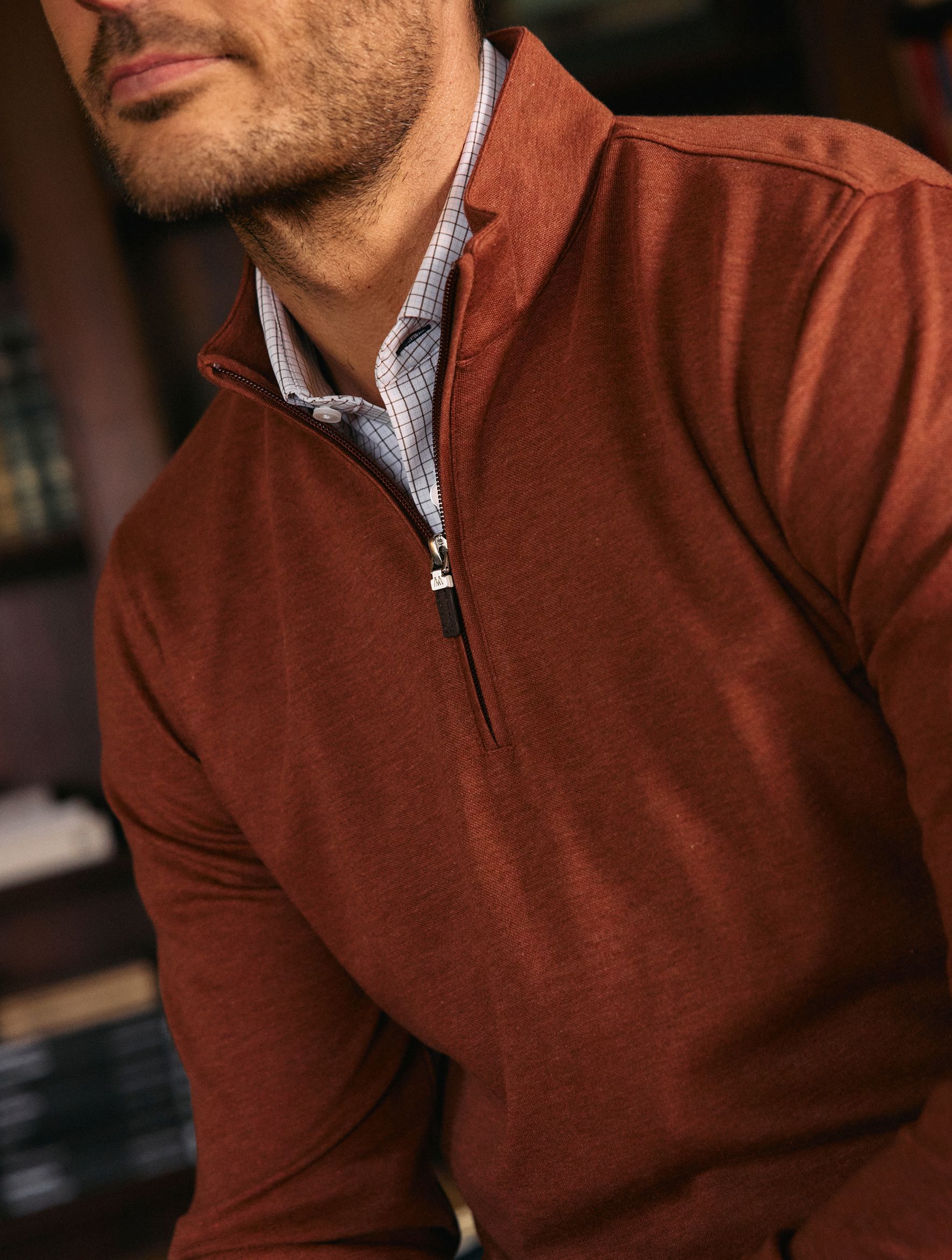 A close-up detail shot of a man wearing a rust-orange quarter-zip sweater layered over a light grid-pattern collared shirt.