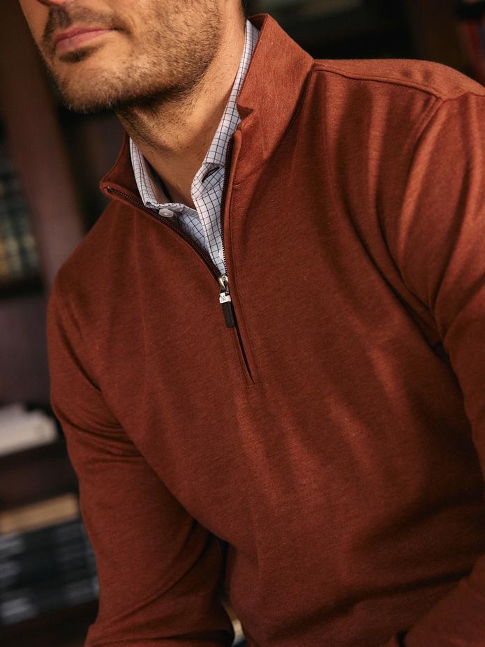 A close-up detail shot of a man wearing a rust-orange quarter-zip sweater layered over a light grid-pattern collared shirt.