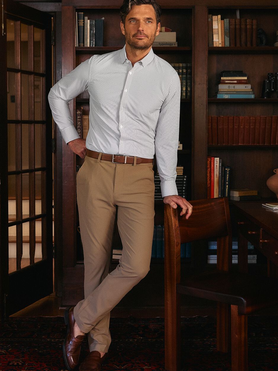A man in a light grey check dress shirt and tan trousers leaning on a wooden chair in a library setting.
