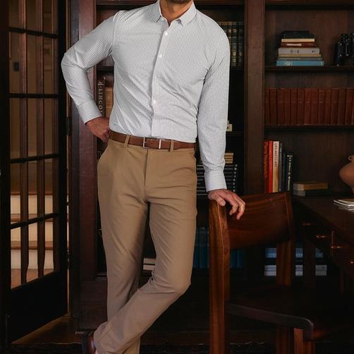 A man in a light grey check dress shirt and tan trousers leaning on a wooden chair in a library setting.