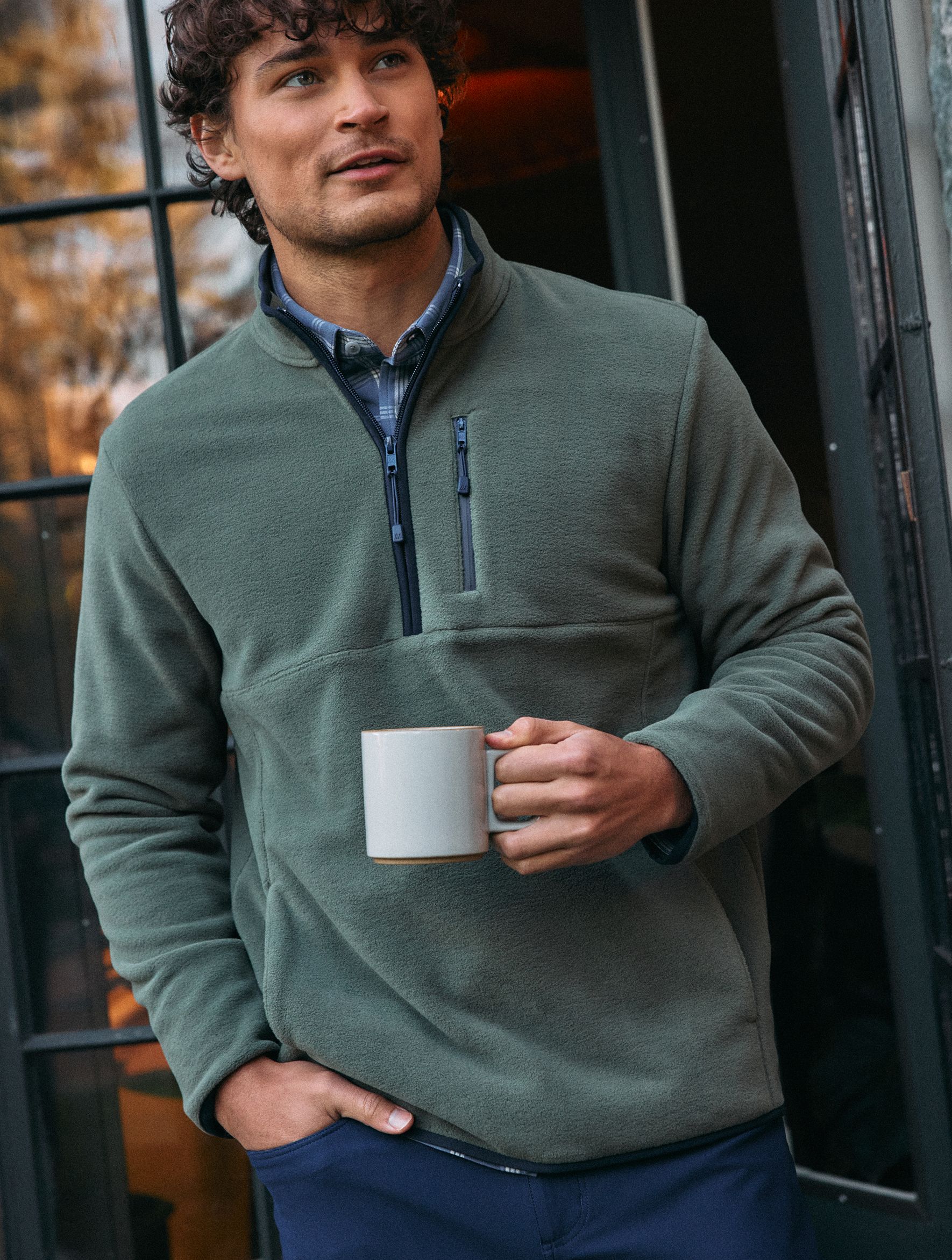 A man wearing an olive green quarter-zip fleece pullover over a plaid shirt holding a coffee mug outdoors.