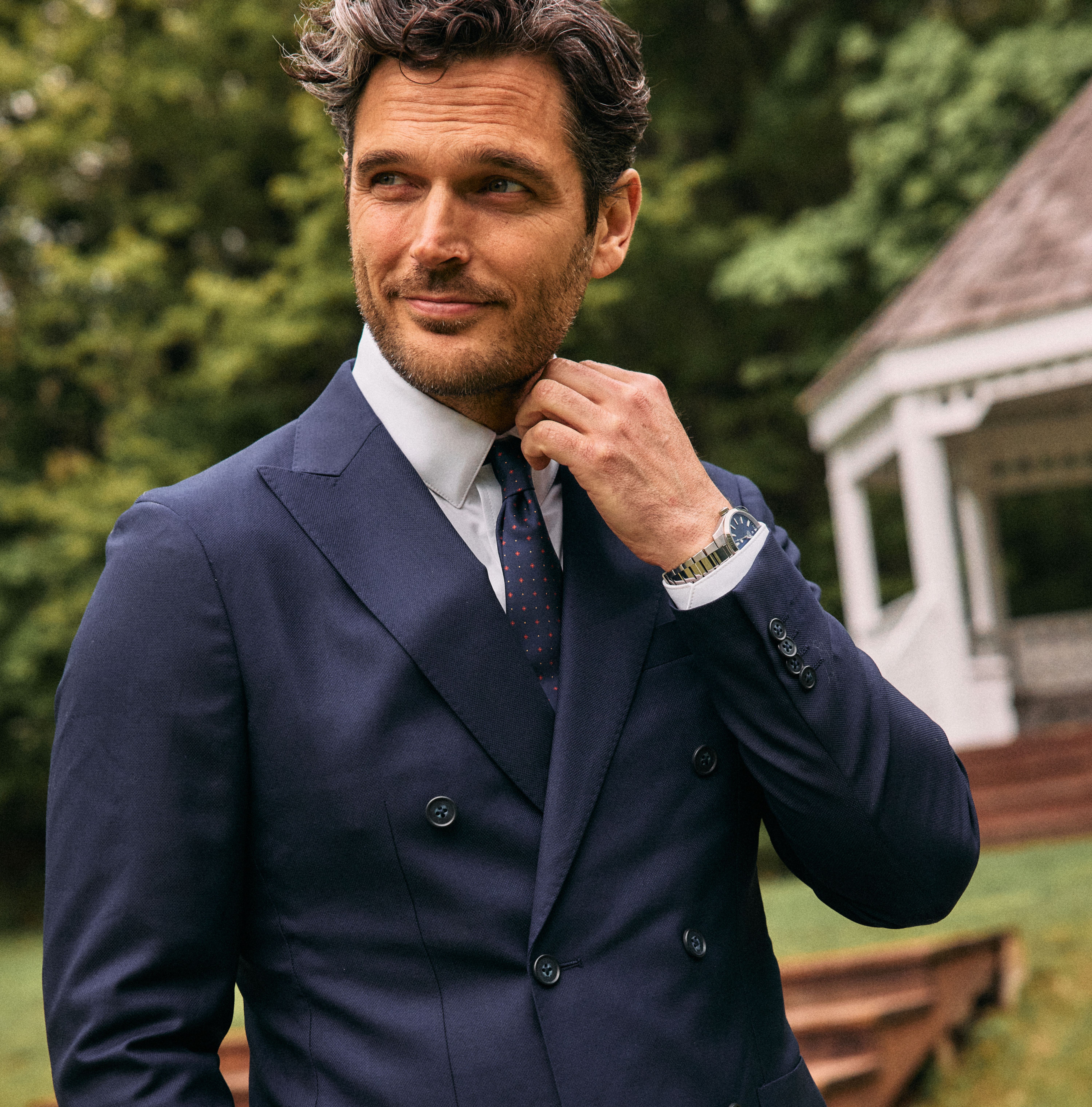 A man adjusting his collar while wearing a double-breasted navy blue suit jacket, white shirt, and patterned red tie outdoors.