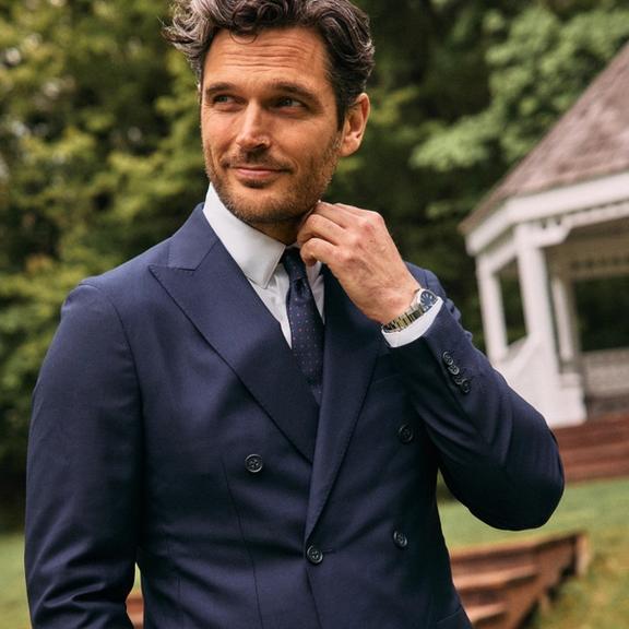 A man adjusting his collar while wearing a double-breasted navy blue suit jacket, white shirt, and patterned red tie outdoors.