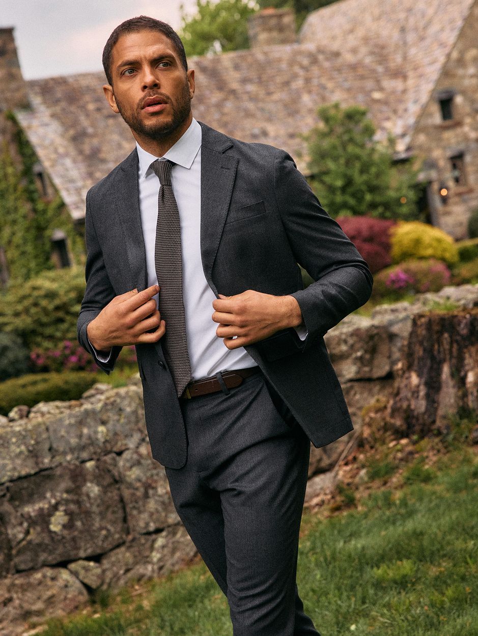 A man adjusting his jacket while wearing a dark charcoal suit, white dress shirt, and knit tie outdoors in a garden setting.