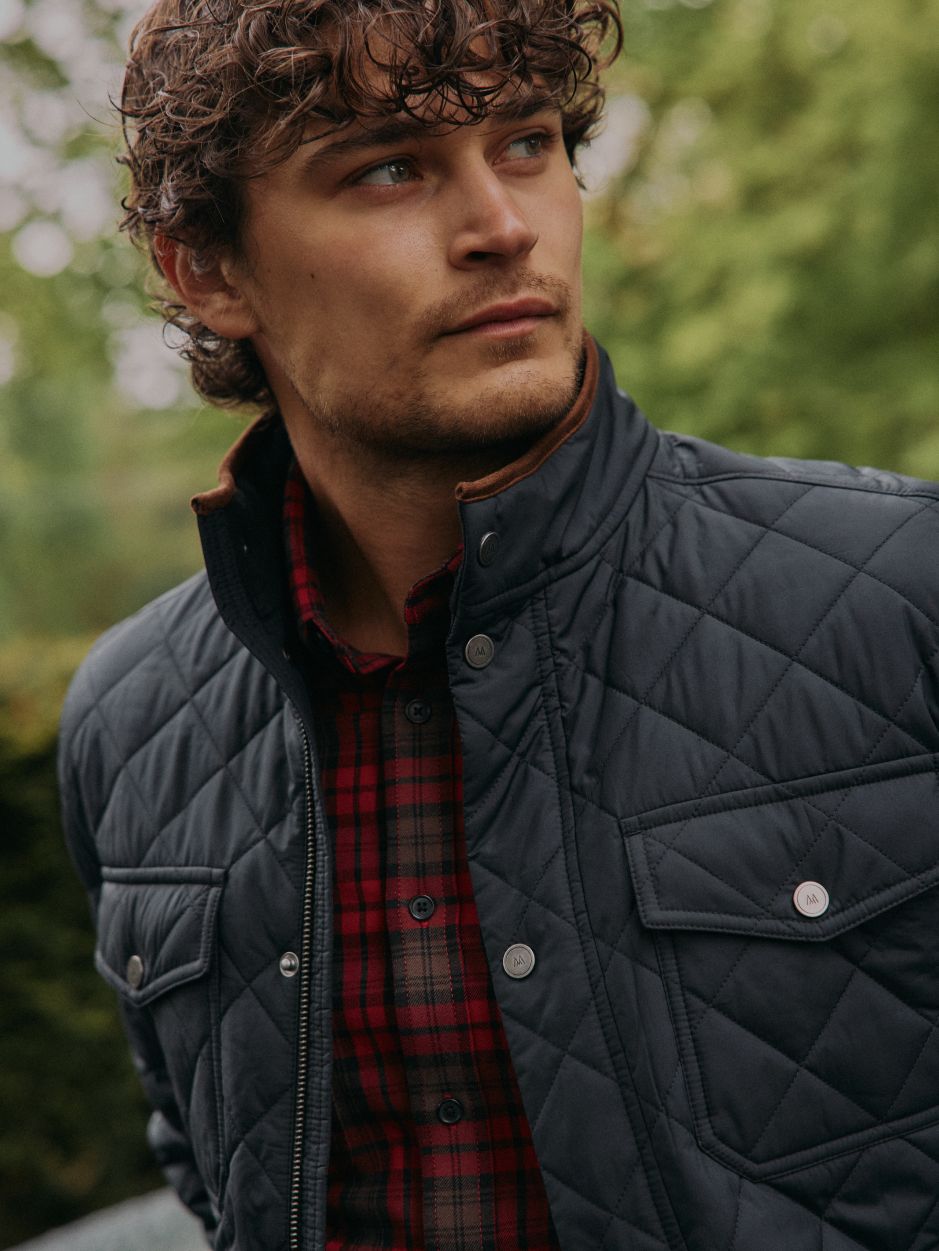 A close-up portrait of a curly-haired man wearing a black quilted jacket over a red plaid button-down shirt.