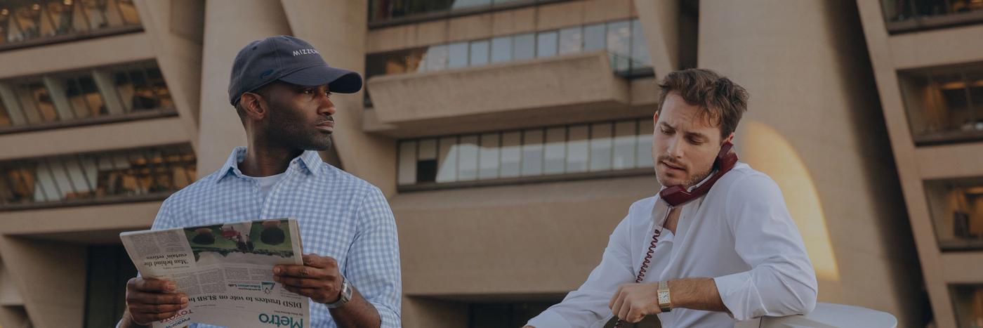 Two men, one talking on a phone, working at a desk outside of a concrete building.
