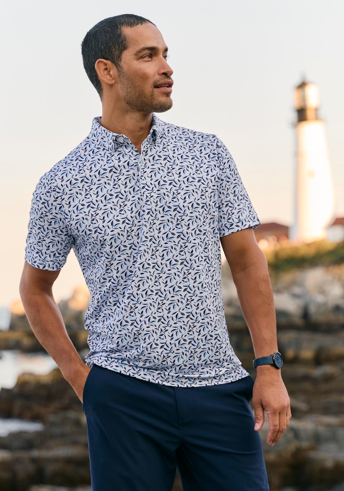 Man in a botanical-print short sleeve shirt and navy shorts on a rocky coast near a lighthouse