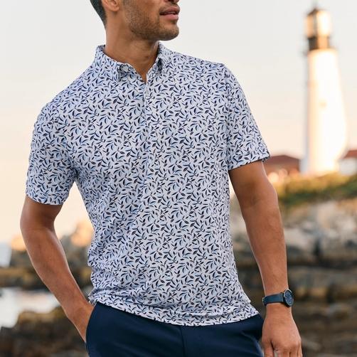 Man in a botanical-print short sleeve shirt and navy shorts on a rocky coast near a lighthouse