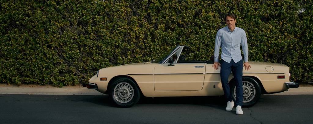 Man in a light blue plaid button-down shirt, navy pants, and white sneakers leaning against a cream vintage convertible in front of a hedge.