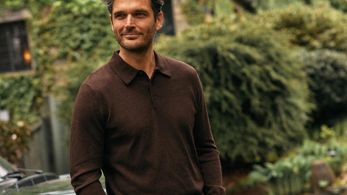 A man wearing a dark brown long-sleeve knit polo shirt posing outdoors with lush greenery in the background.