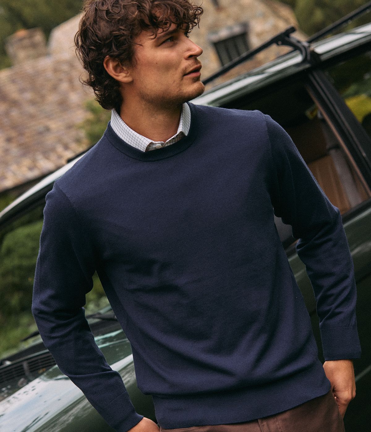 Man in a navy Heritage Crewneck Sweater over a white check collared shirt, standing beside a vintage green car.