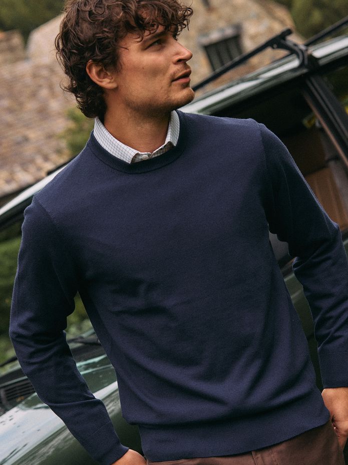 Man in a navy Heritage Crewneck Sweater over a white check collared shirt, standing beside a vintage green car.
