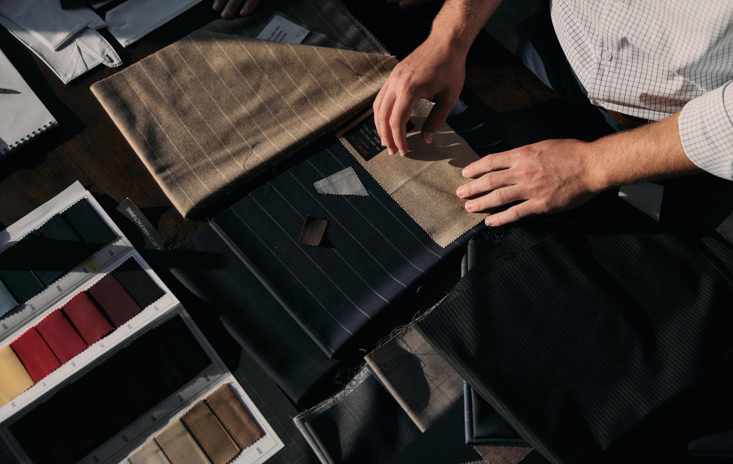 An overhead shot of hands examining fabric swatches and textiles arranged on a dark wooden table alongside color sample cards.