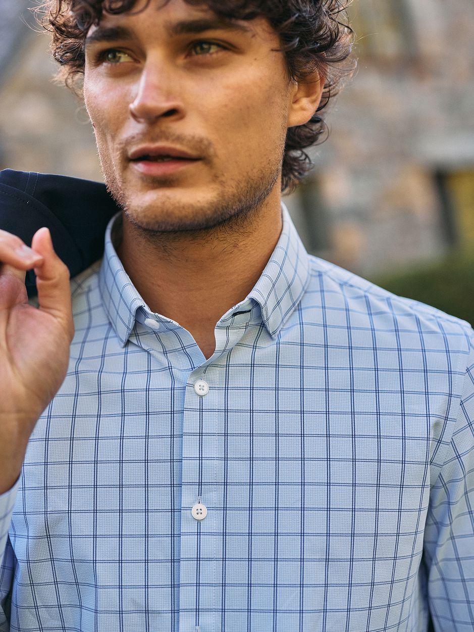 A close-up shot of a curly-haired man outdoors wearing a light blue grid-pattern collared dress shirt with a jacket draped over his shoulder.