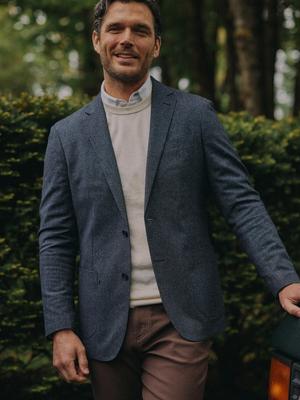 A man smiling while standing next to a car outdoors, wearing a grey textured blazer, an off-white crewneck sweater, a collared shirt, and brown trousers.