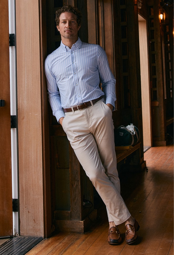 Man in a light blue striped shirt and stone Prescott pants leaning in a wood-paneled hallway