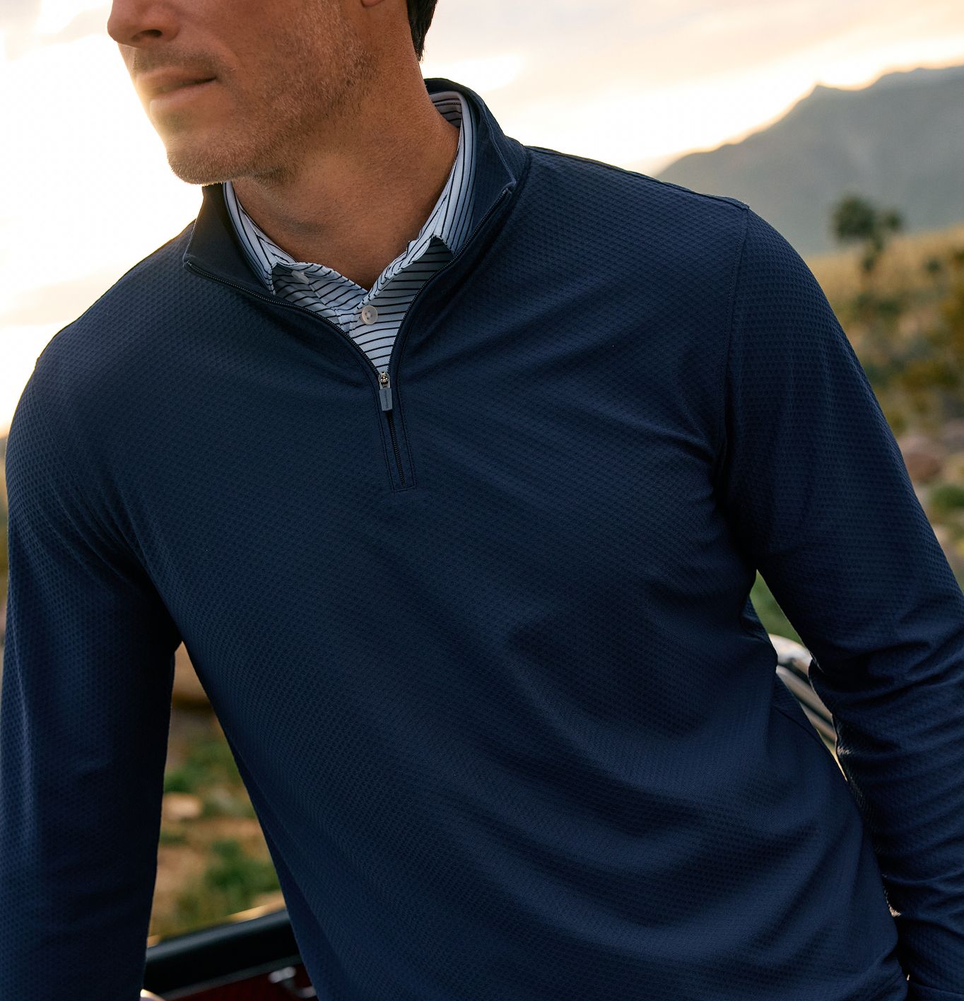 Close-up of a man wearing a navy quarter-zip pullover layered over a blue and white striped collared shirt, outdoors at sunset with mountains in the background.