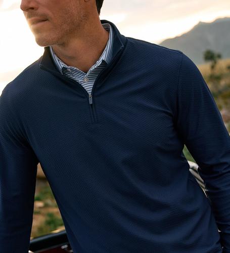 Close-up of a man wearing a navy quarter-zip pullover layered over a blue and white striped collared shirt, outdoors at sunset with mountains in the background.