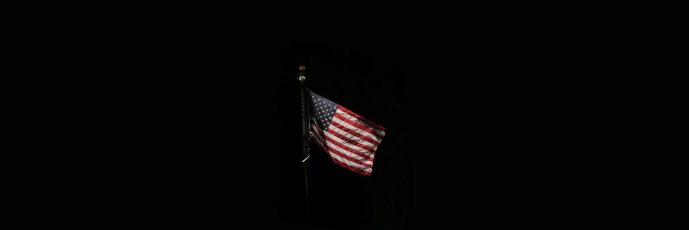 The US flag, flying on a flagpole against a black background.
