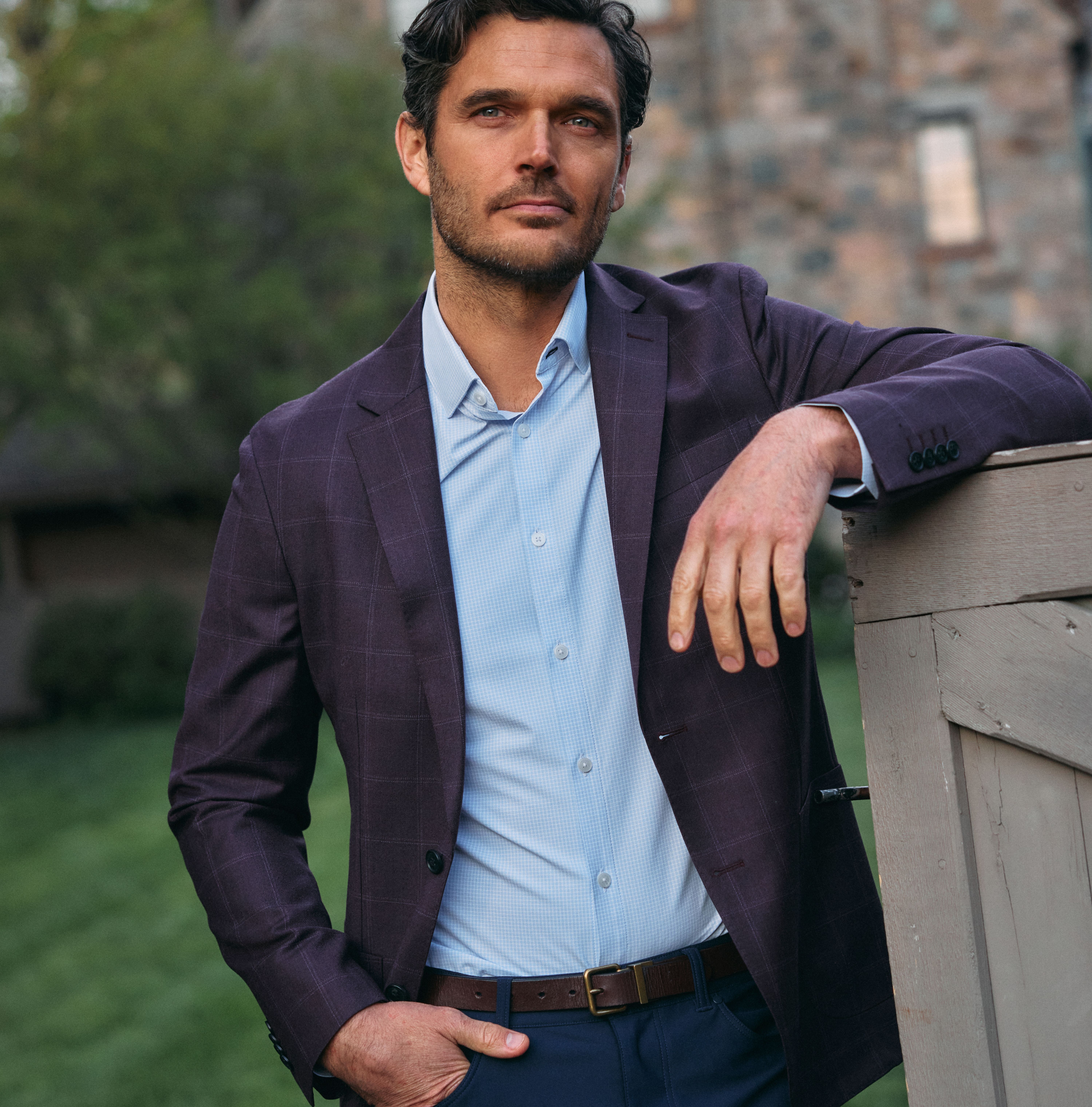 A man with dark wavy hair leaning on a wooden fence, wearing a plum windowpane blazer over a light blue collared shirt and navy trousers.