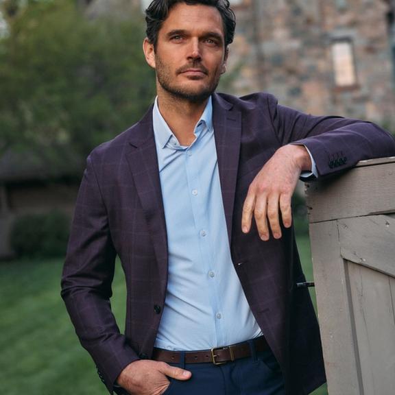 A man with dark wavy hair leaning on a wooden fence, wearing a plum windowpane blazer over a light blue collared shirt and navy trousers.