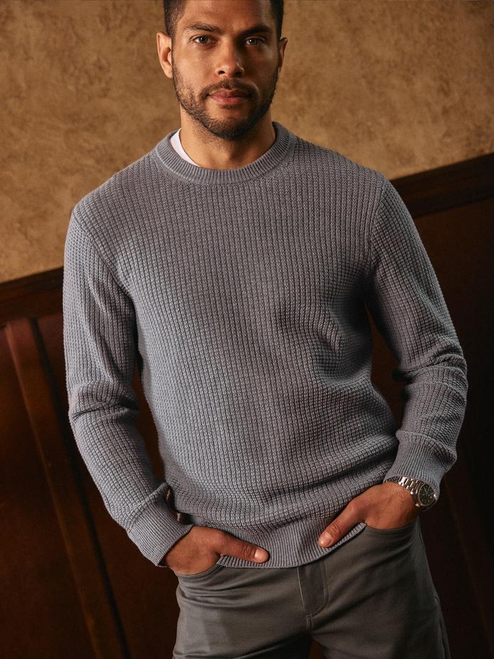 A man in a grey textured waffle-knit crewneck sweater and dark pants posing indoors.