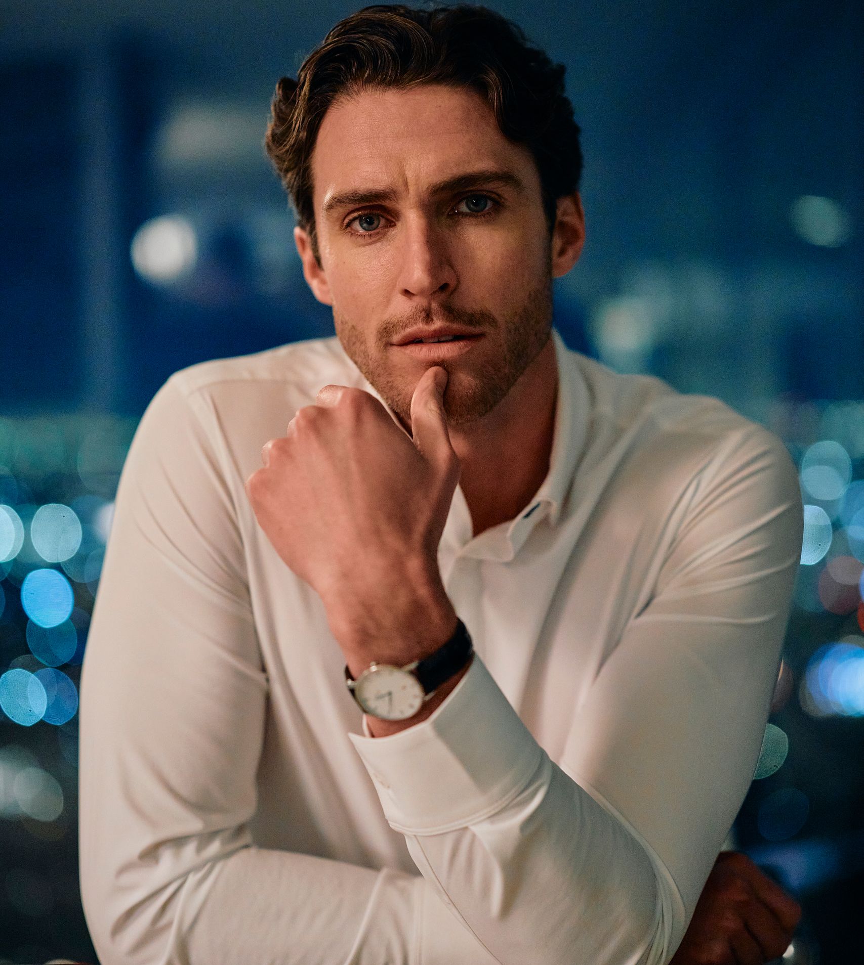 A man in a crisp white dress shirt rests his chin on his hand, city lights glowing behind him.