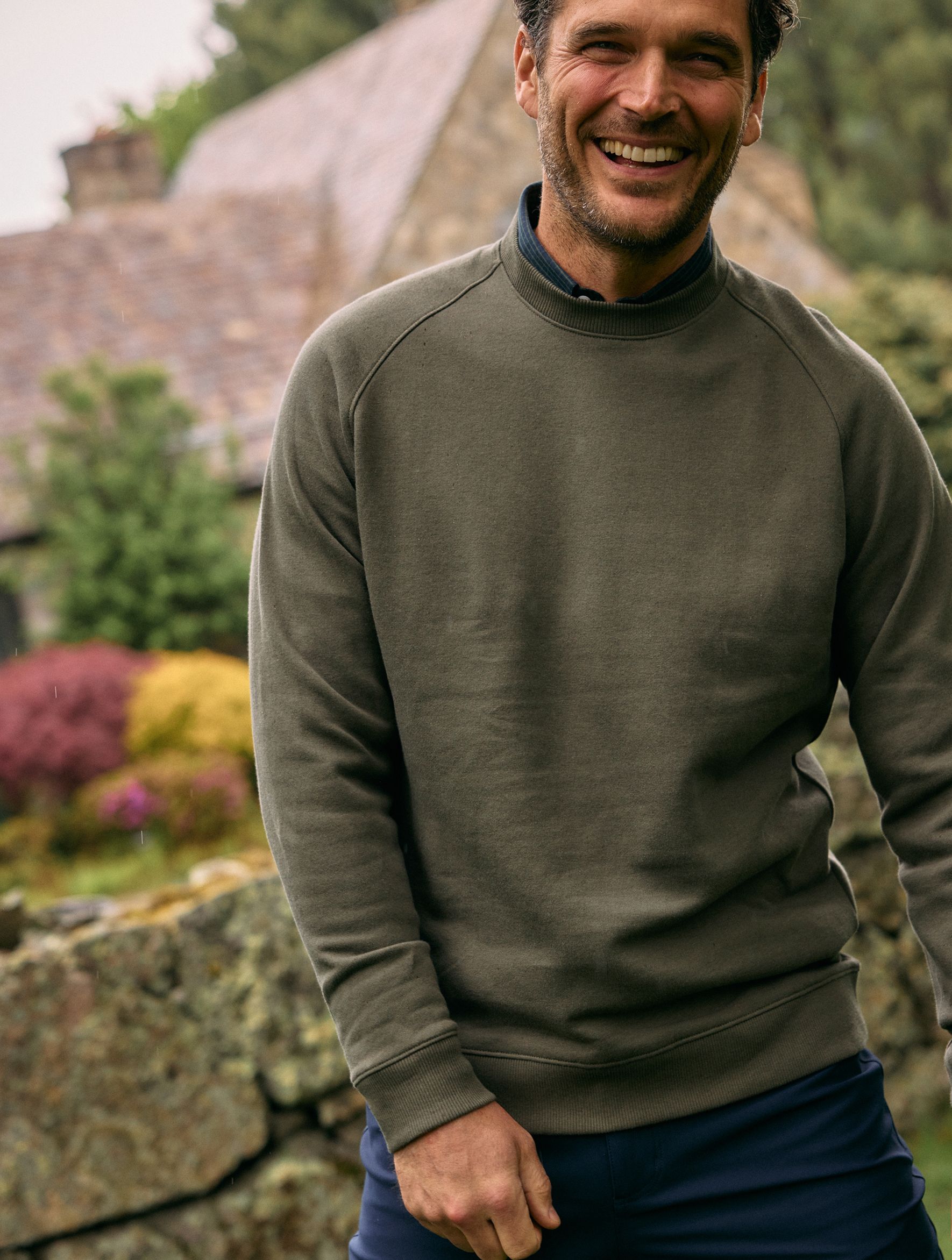 A smiling man wearing an olive green raglan crewneck sweatshirt over a dark shirt standing outdoors in front of a stone wall.