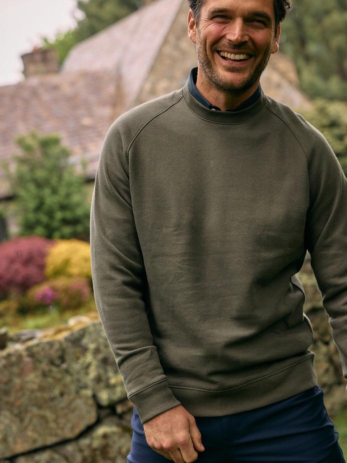 A smiling man wearing an olive green raglan crewneck sweatshirt over a dark shirt standing outdoors in front of a stone wall.