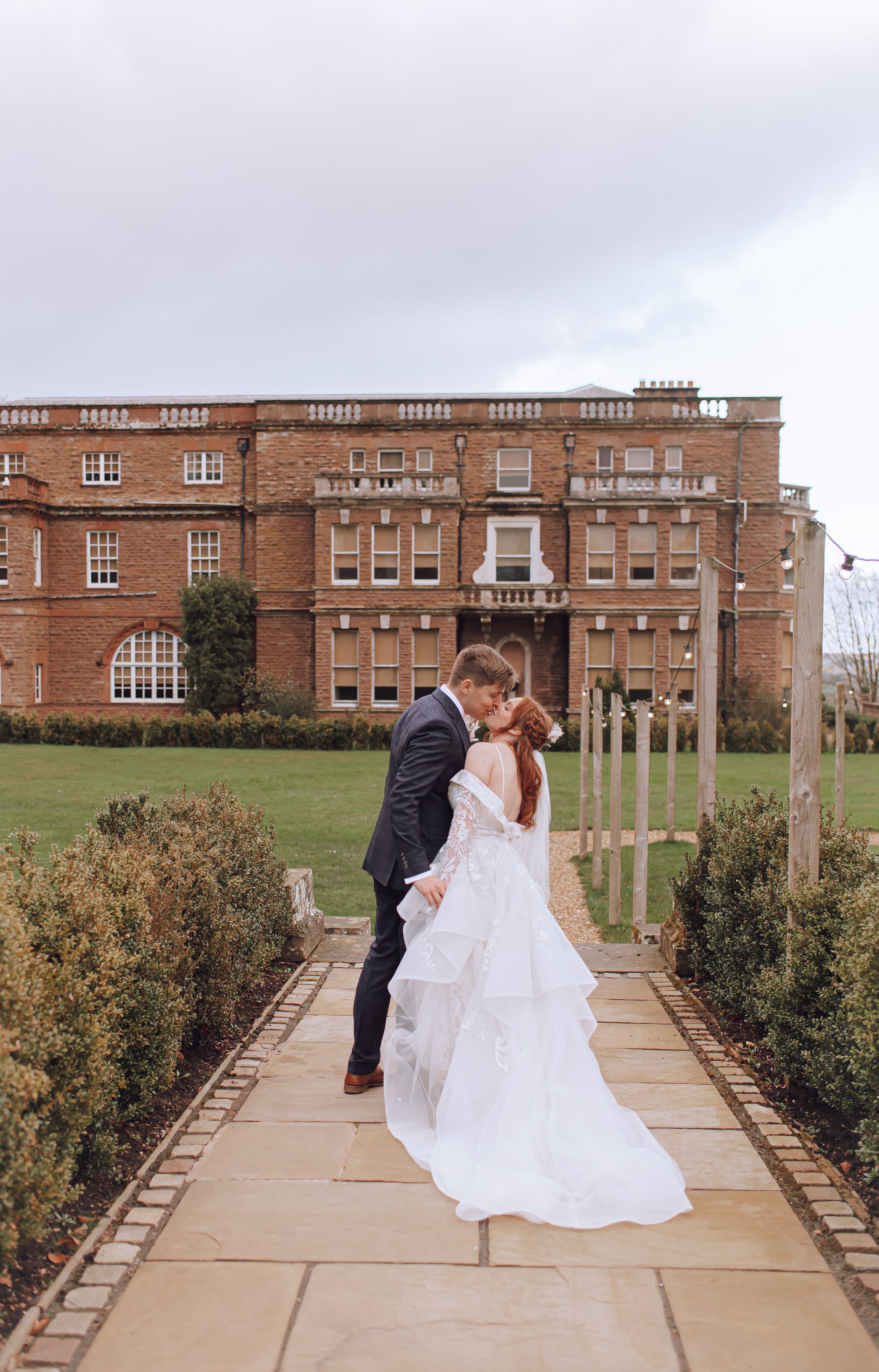 A bride and groom share an intimate moment outside a large, historic brick mansion. The groom is wearing a dark suit, and the bride is in an elaborate white wedding gown with lace details. They are standing close together on a paved pathway, surrounded by neatly trimmed hedges and a well-maintained lawn. The sky is overcast, adding a soft, romantic ambiance to the scene.