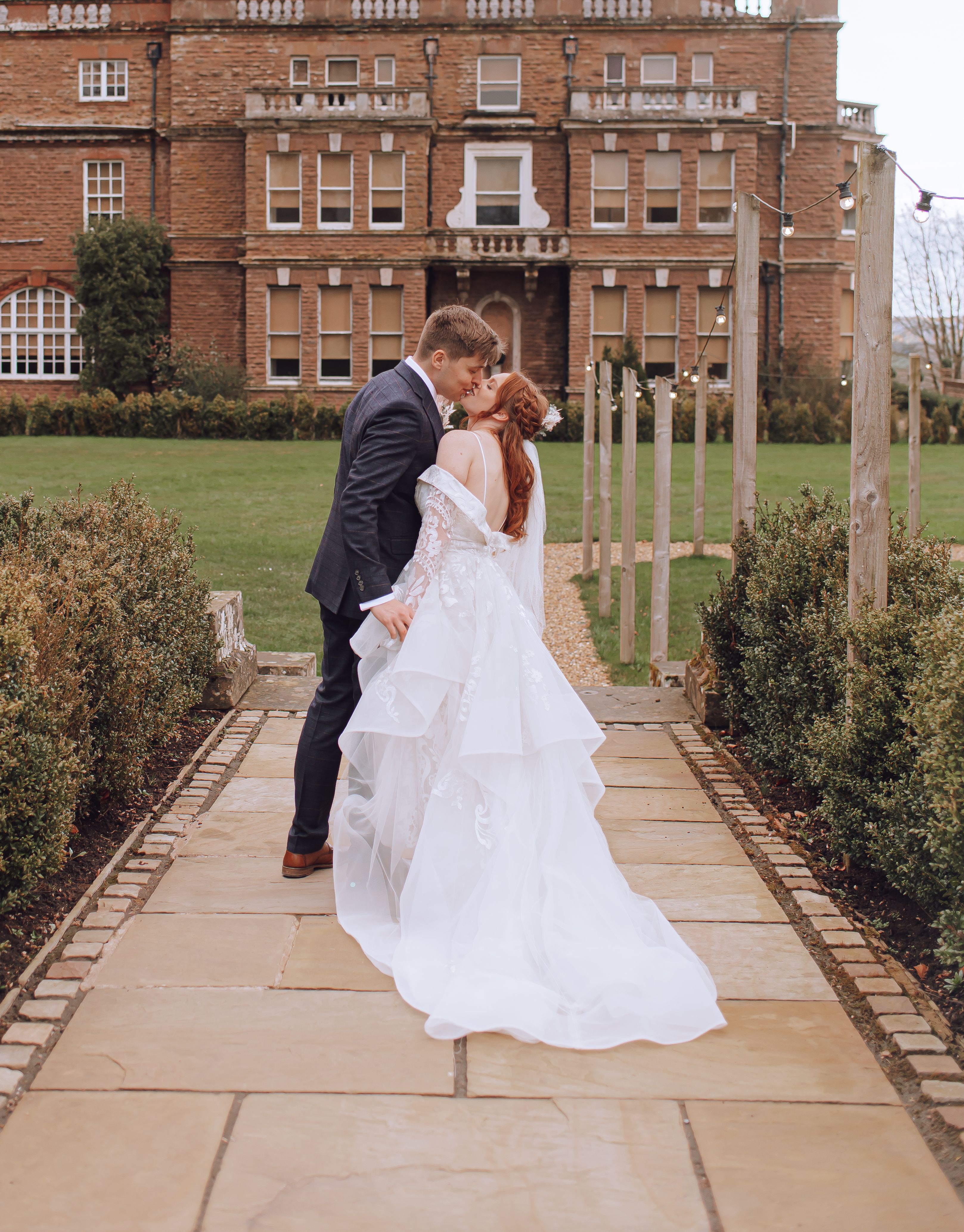 A bride and groom share an intimate moment outside a large, historic brick mansion. The groom is wearing a dark suit, and the bride is in an elaborate white wedding gown with lace details. They are standing close together on a paved pathway, surrounded by neatly trimmed hedges and a well-maintained lawn. The sky is overcast, adding a soft, romantic ambiance to the scene.