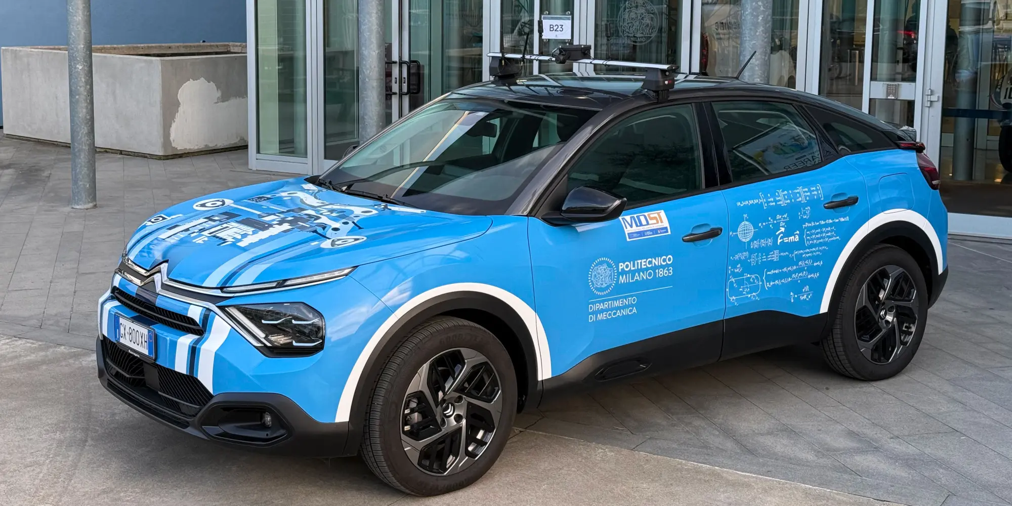 Auto di ricerca blu con sensori sul tetto e grafiche tecniche, parcheggiata davanti a un edificio.