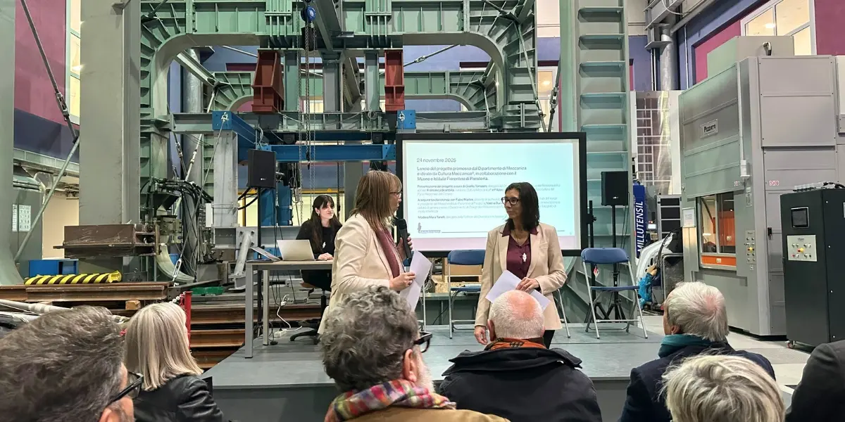 Due relatrici sul palco di un laboratorio industriale parlano davanti a uno schermo con il programma dell’evento, mentre una persona lavora al computer accanto a loro.