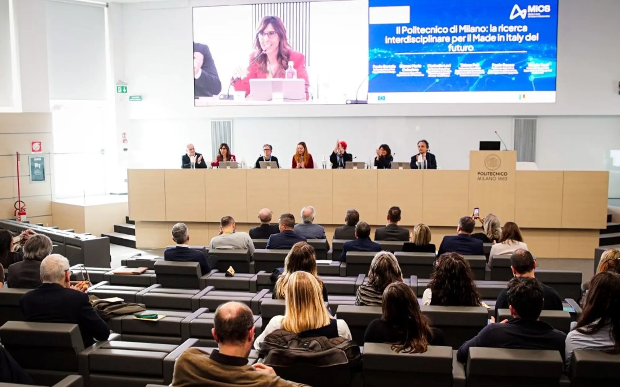 Pubblico assiste a una conferenza con relatori sul palco e presentazione su grande schermo.