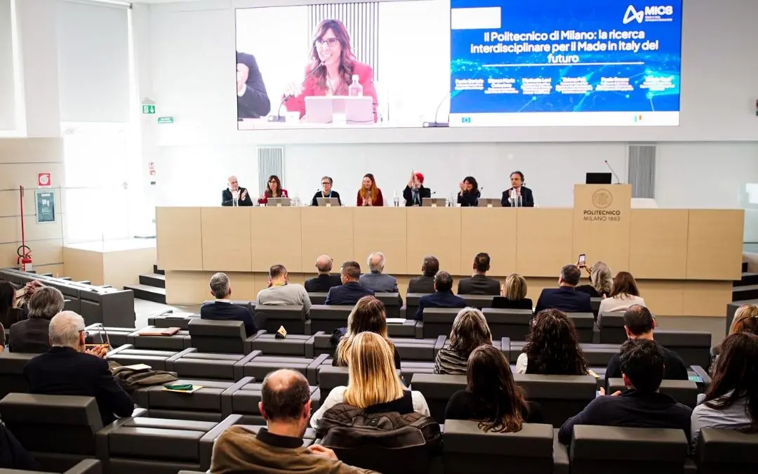 Pubblico assiste a una conferenza con relatori sul palco e presentazione su grande schermo.
