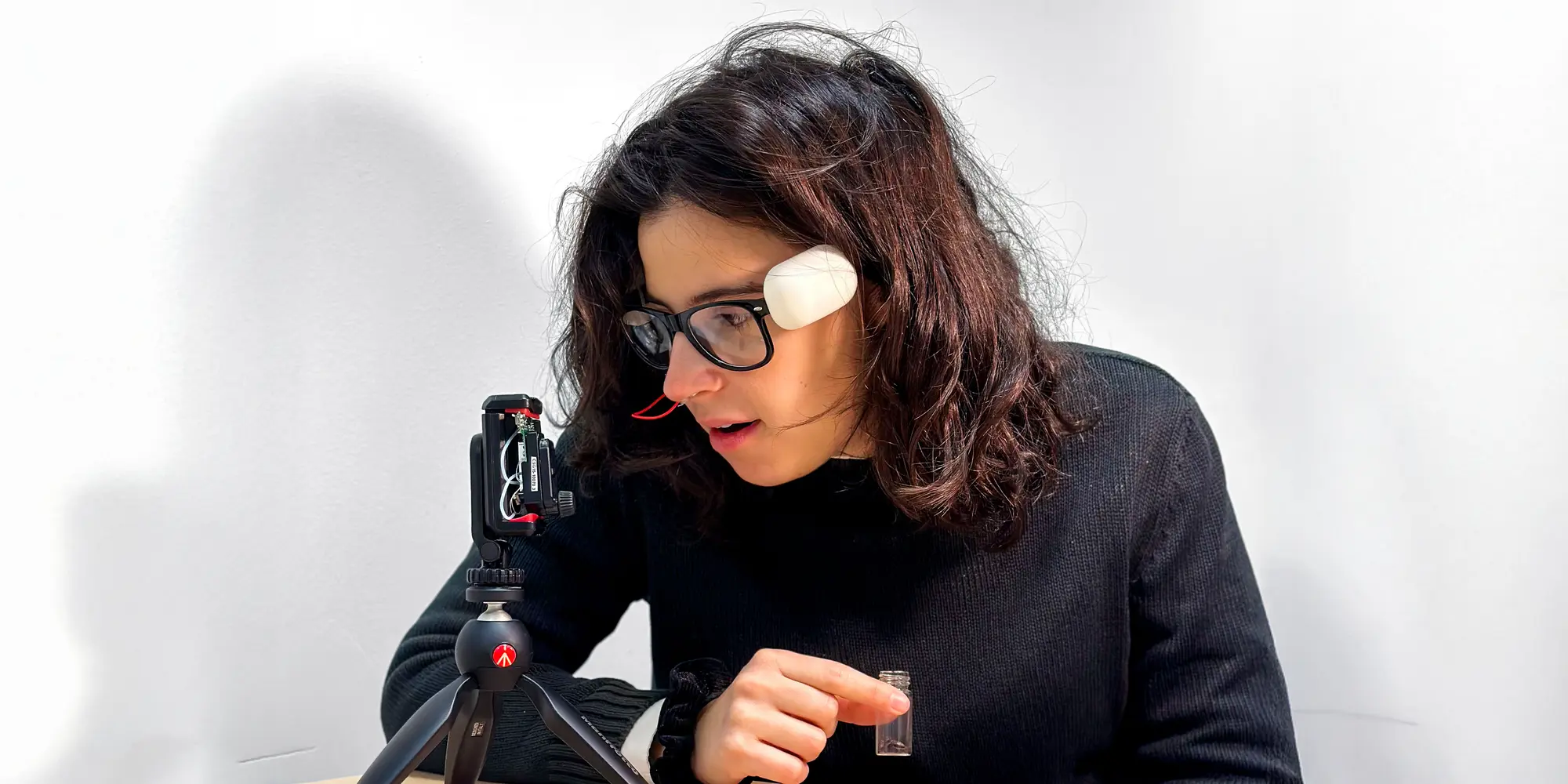 Person pointing at a small container while observing a technological device on a tripod, wearing a facial sensor.