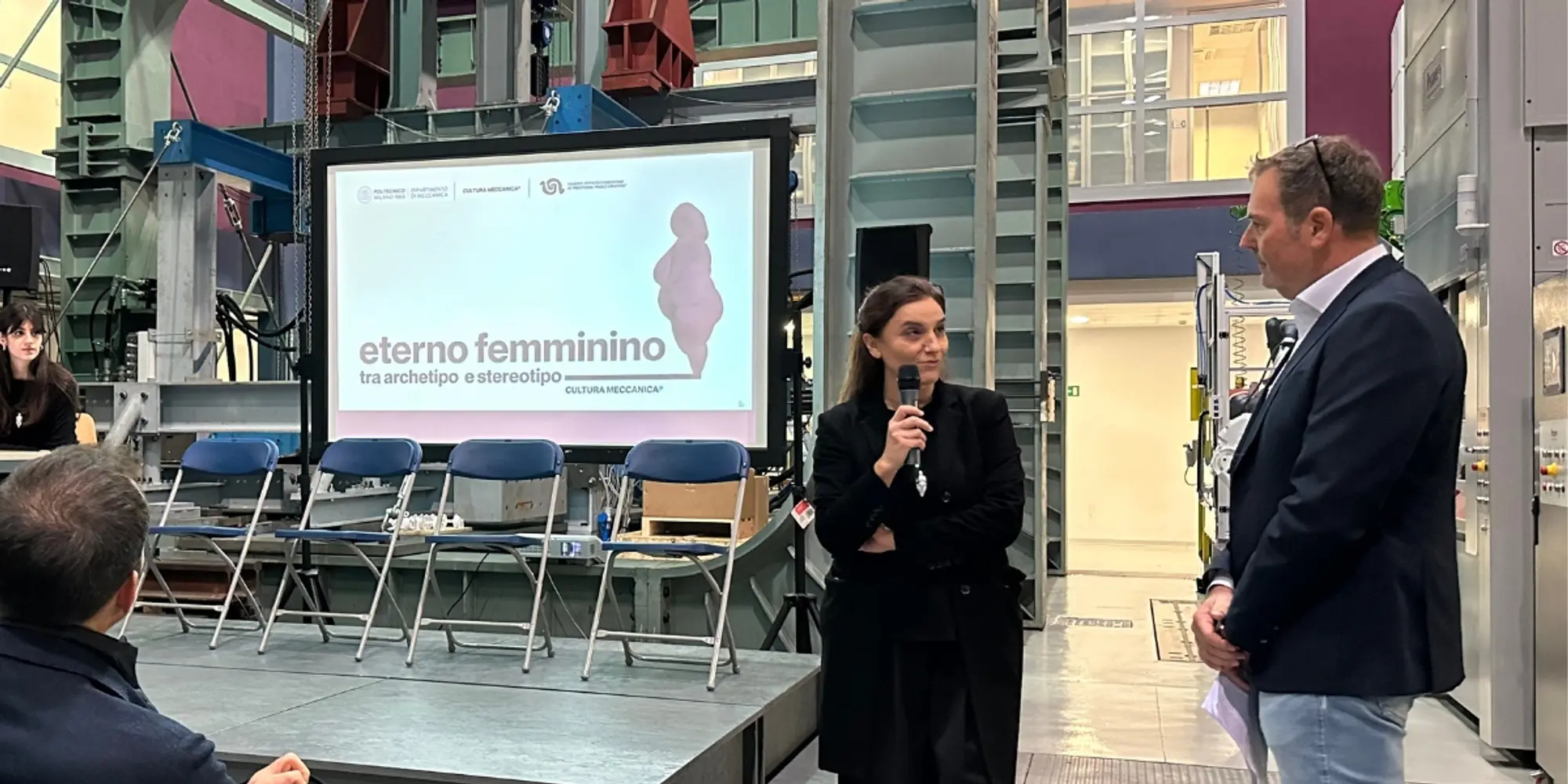 Due persone parlano con microfono davanti a uno schermo con il titolo “Eterno femminino – tra archetipo e stereotipo” in un laboratorio industriale.